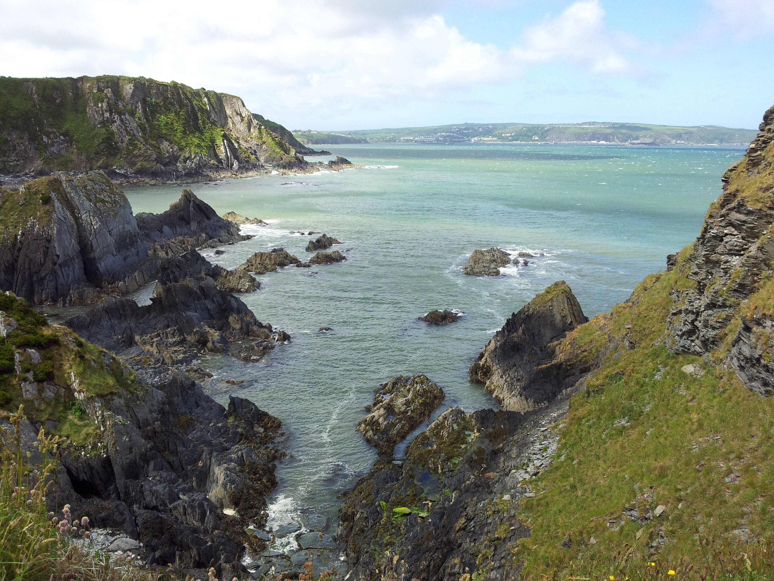 A coastal landscape with rocky cliffs, a narrow waterway, and distant hills.