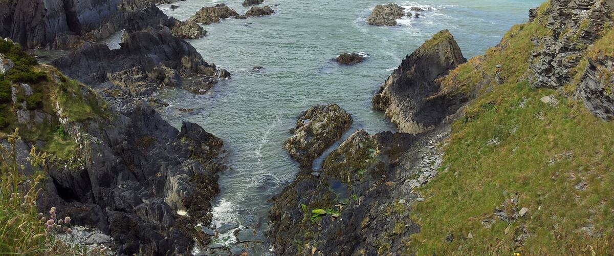 A coastal landscape with rocky cliffs, a narrow waterway, and distant hills.