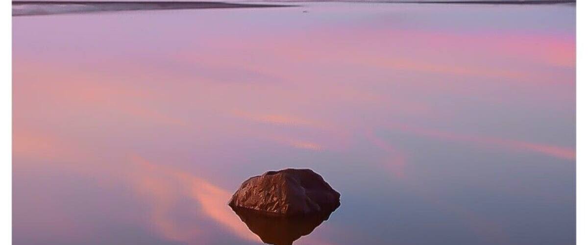 A calm body of water with a single rock in the foreground and a clear sky.