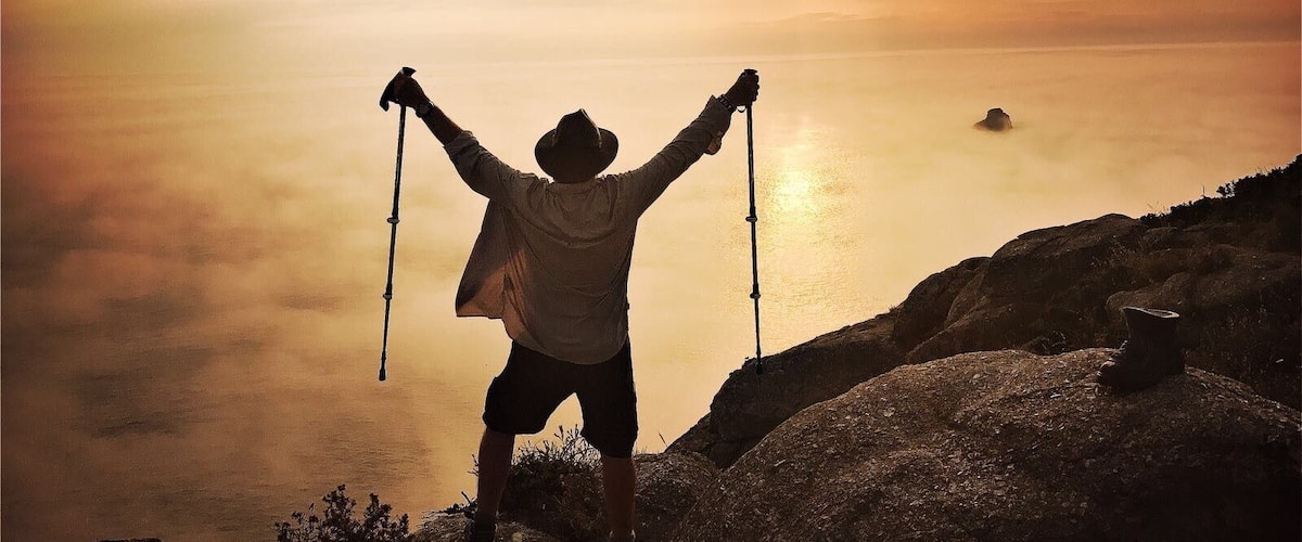 A person hiking with a backpack and hiking poles, standing on a rocky cliff overlooking a body of water during sunset.
