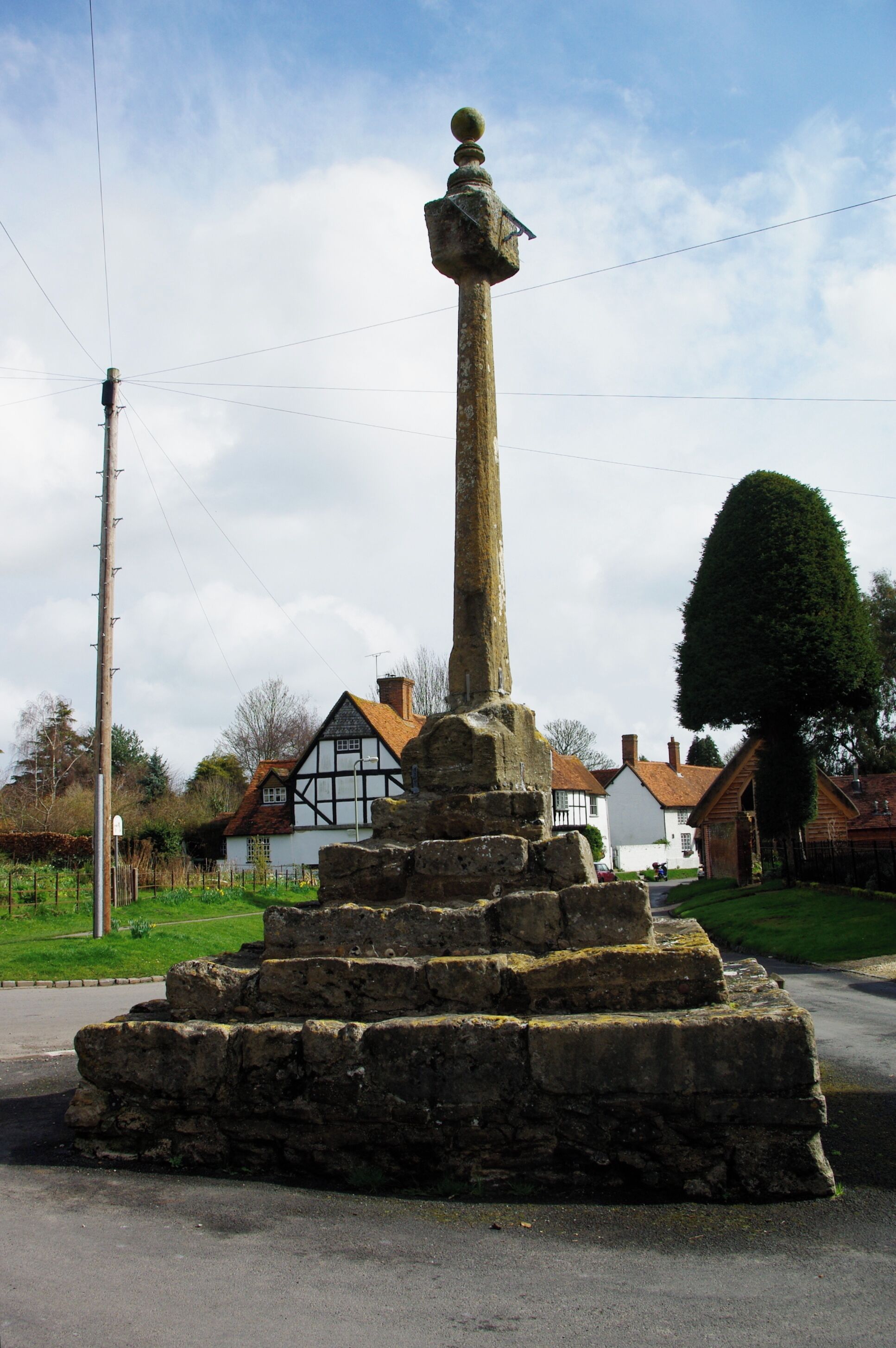 A stone monument with a cross on top, surrounded by a stone base, in a rural setting with houses and a clear sky.