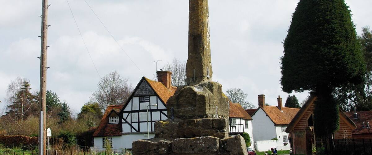 A stone monument with a cross on top, surrounded by a stone base, in a rural setting with houses and a clear sky.