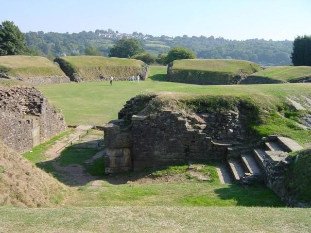 Ruinas antiguas con muros y escalones de piedra, rodeadas de campos verdes y colinas lejanas.