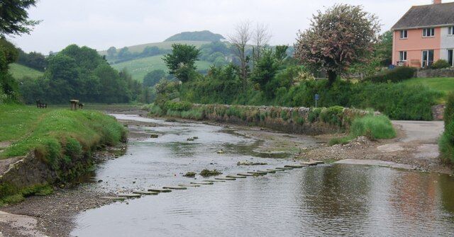 A calm river with a stone bridge, lush greenery, and a pink house in the background.