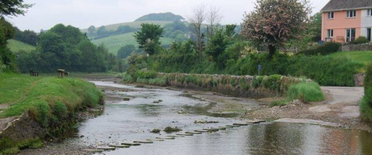 A calm river with a stone bridge, lush greenery, and a pink house in the background.