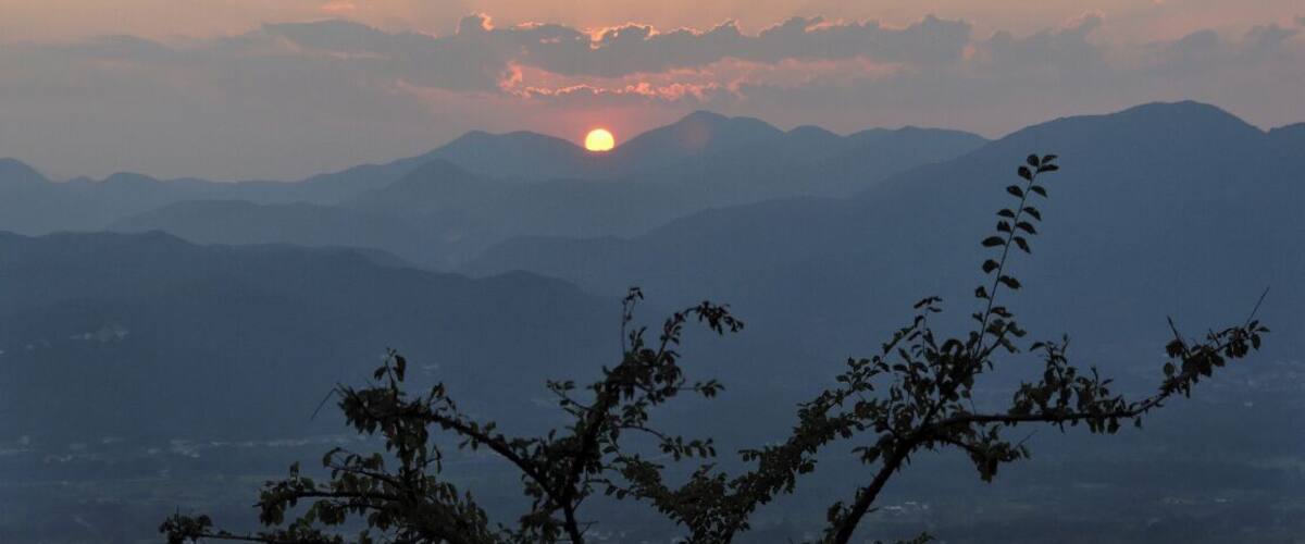 A sunset over a mountain range with the sun partially obscured by clouds.