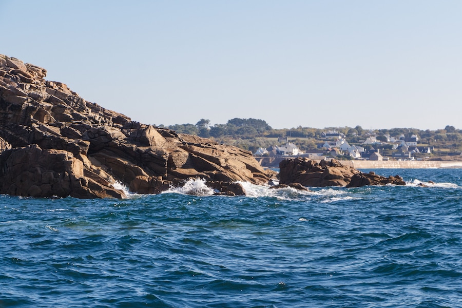 A rocky coastline with waves crashing against the rocks and a town in the background.