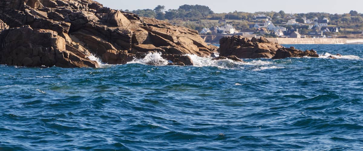 A rocky coastline with waves crashing against the rocks and a town in the background.
