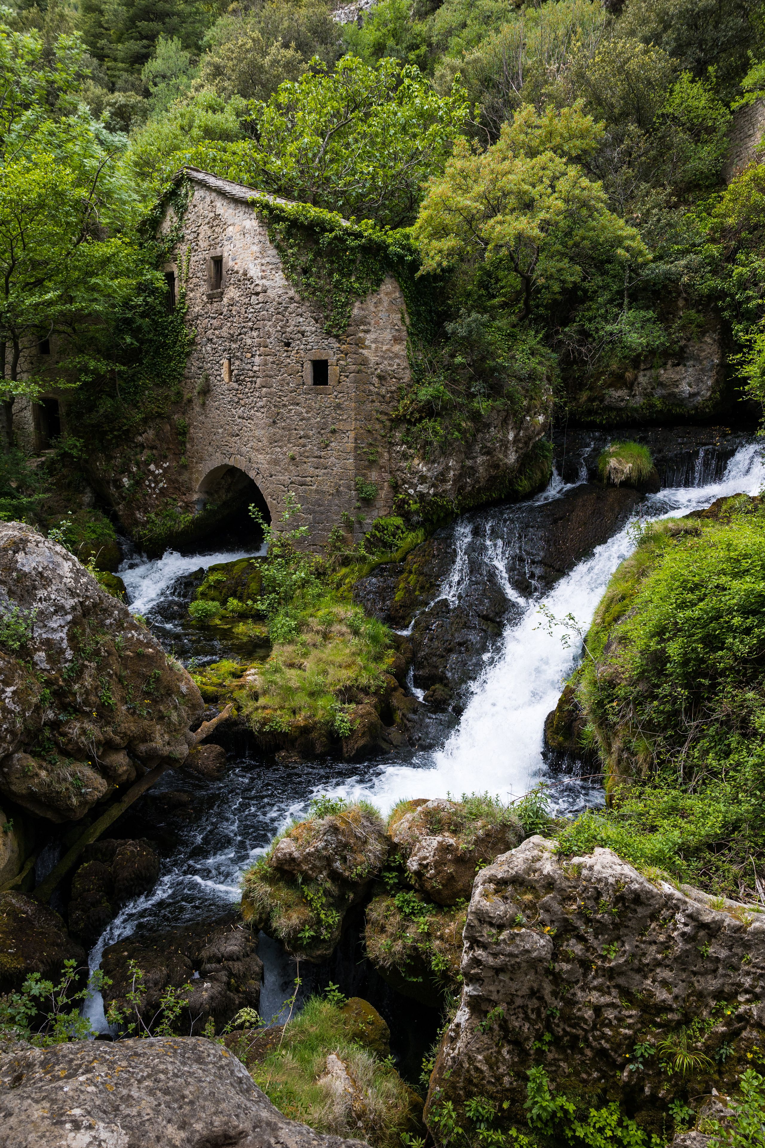 A stone bridge over a waterfall, surrounded by lush greenery and a rocky landscape.
