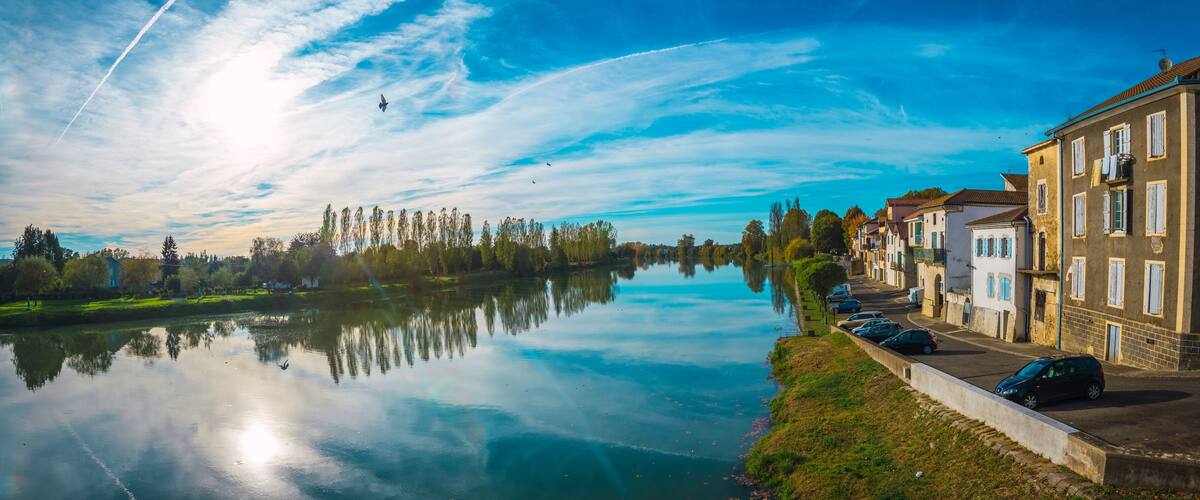 A calm river reflecting buildings and trees, with a clear sky and a parked car along the riverbank.