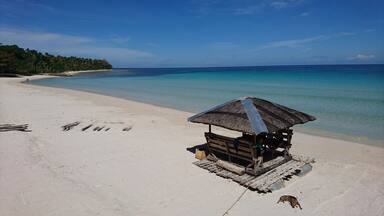 A beach with a wooden hut and a thatched roof, surrounded by clear turquoise water and a sandy shore.