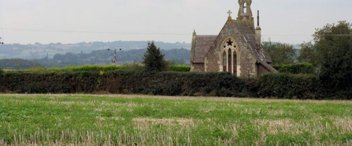A small church with a pointed steeple and a cross on top, surrounded by a field and hedgerows.