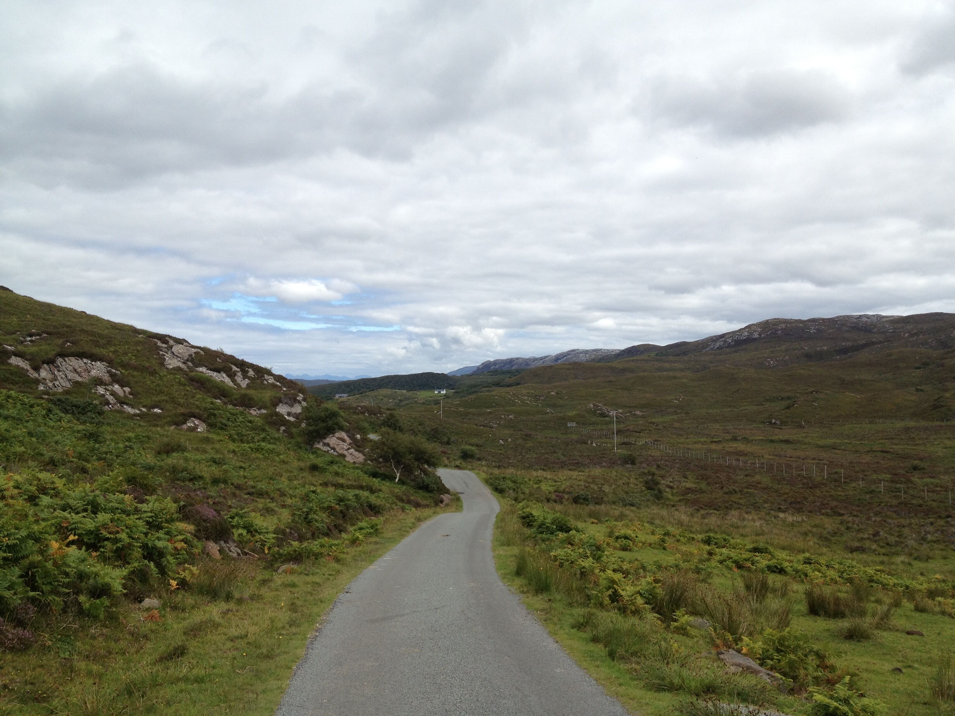 A narrow road winding through a hilly landscape with sparse vegetation and distant mountains under a cloudy sky.