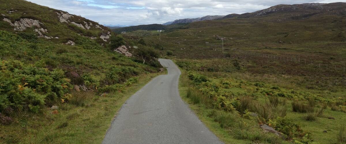A narrow road winding through a hilly landscape with sparse vegetation and distant mountains under a cloudy sky.