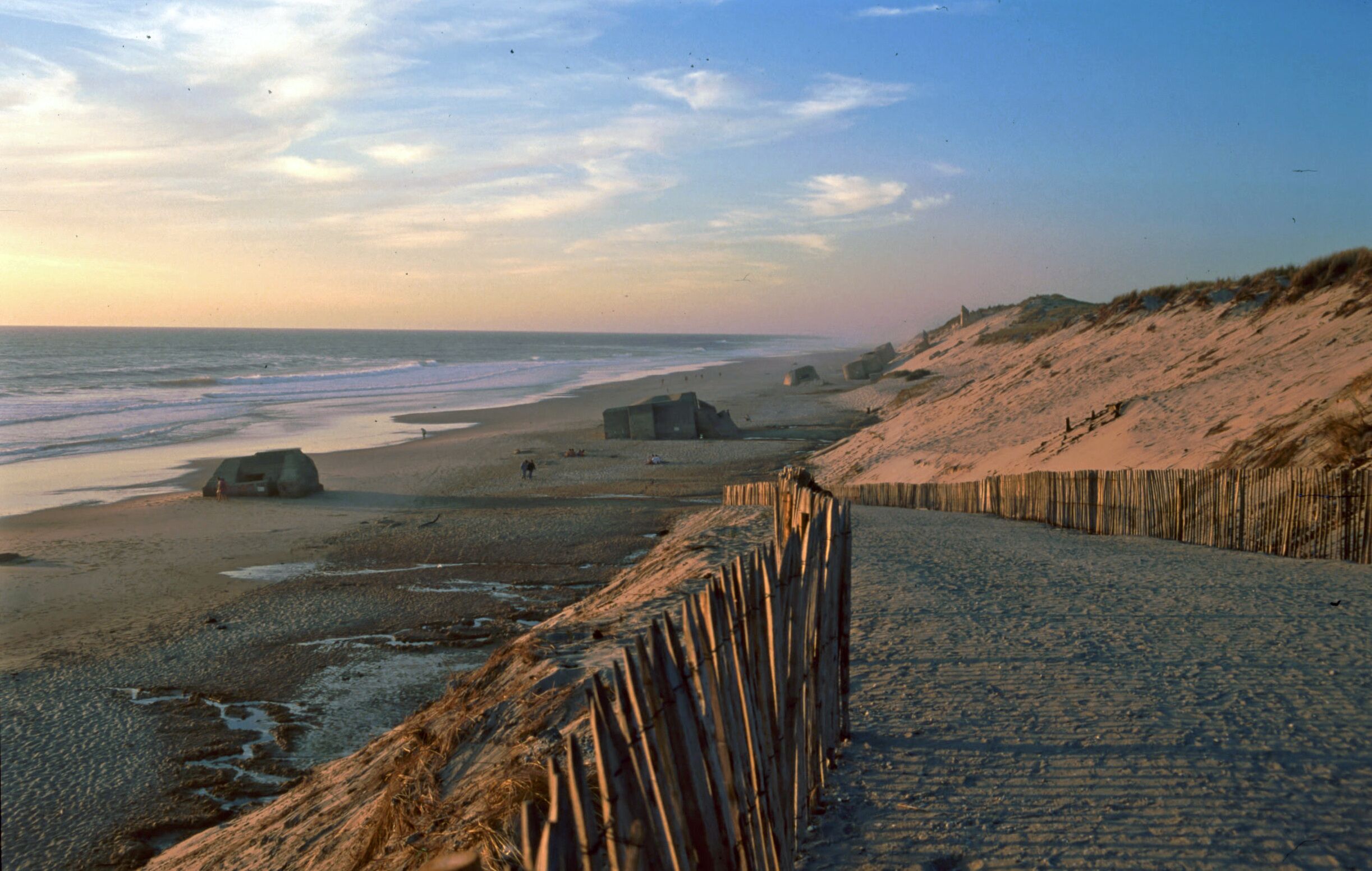A beach with a wooden fence, a dune, and a small structure near the water.