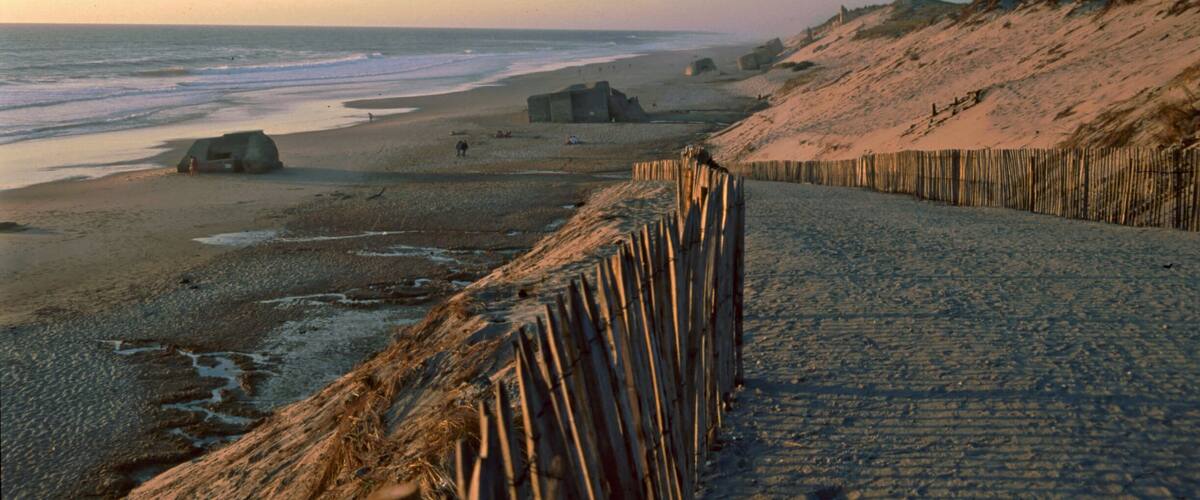 A beach with a wooden fence, a dune, and a small structure near the water.