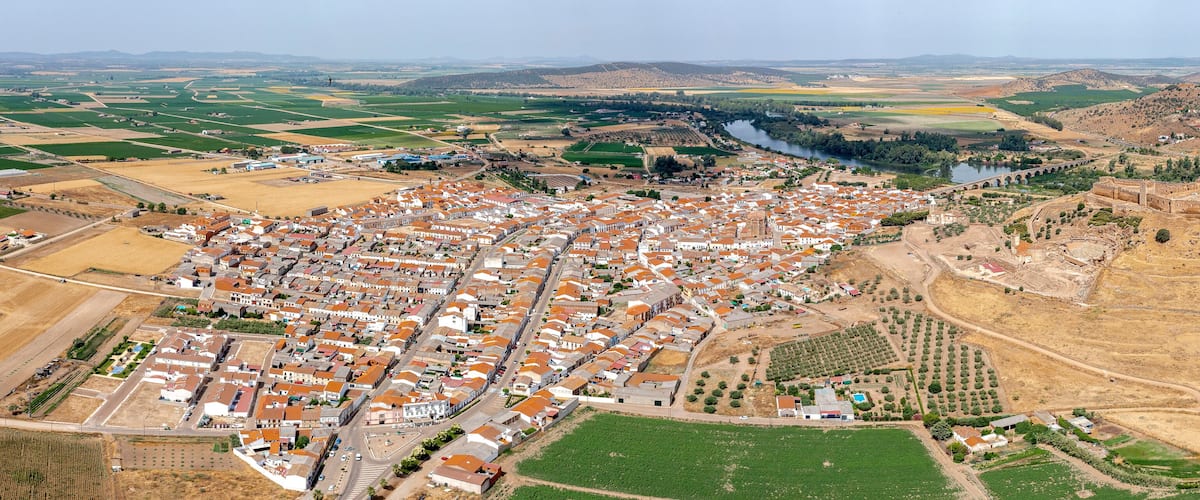 An aerial view of a town with a river, agricultural fields, and a large hill with ruins.