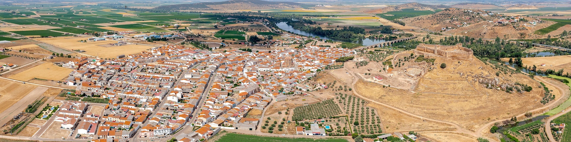 An aerial view of a town with a river, agricultural fields, and a large hill with ruins.
