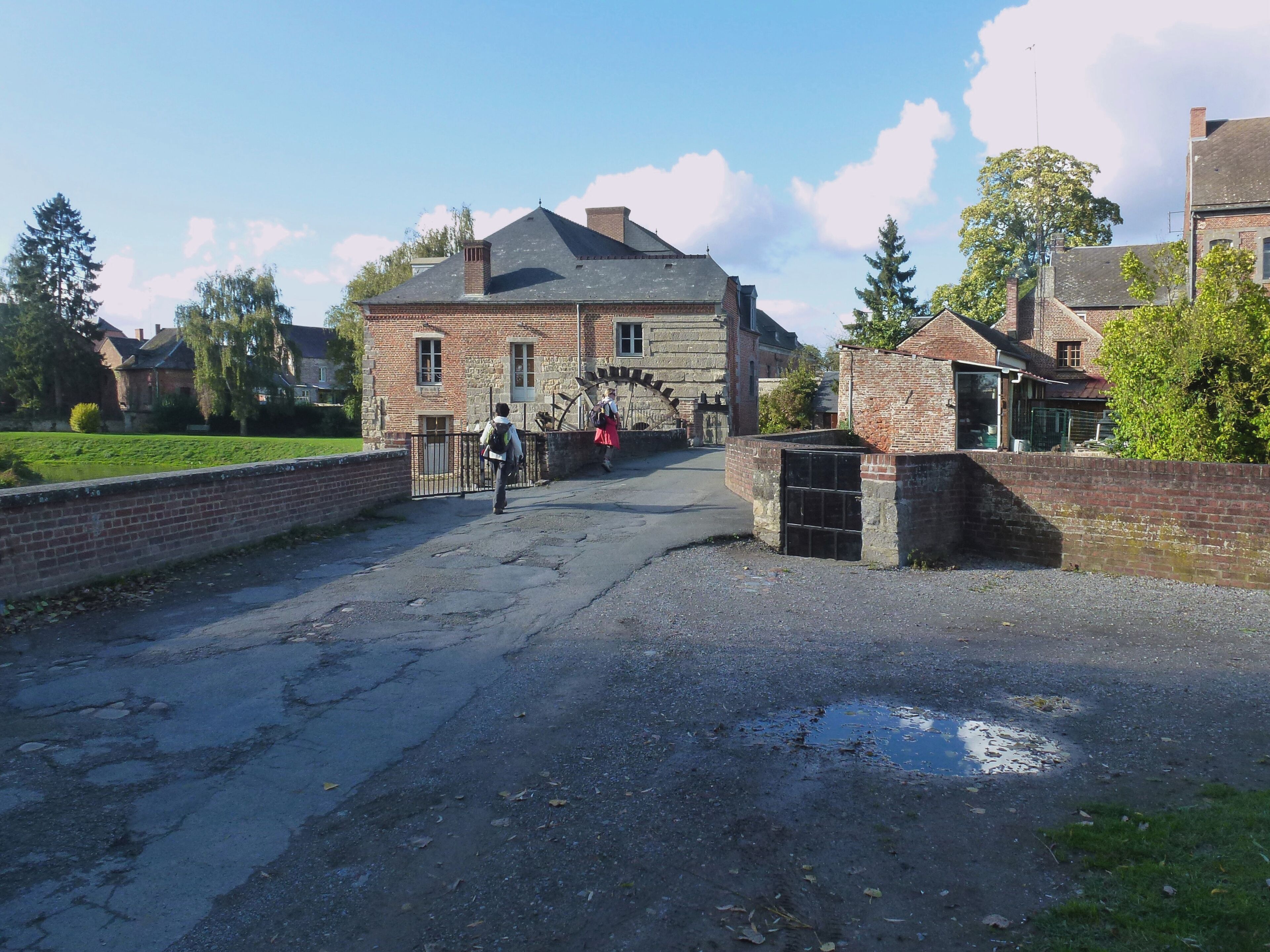 A rural road leading to a stone building with a traditional roof, surrounded by greenery and trees.