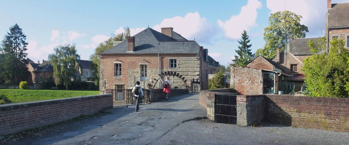A rural road leading to a stone building with a traditional roof, surrounded by greenery and trees.