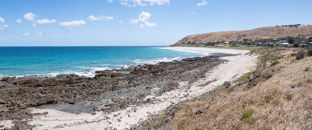 A rocky beach with clear blue water, a sandy shore, and a hillside with houses.