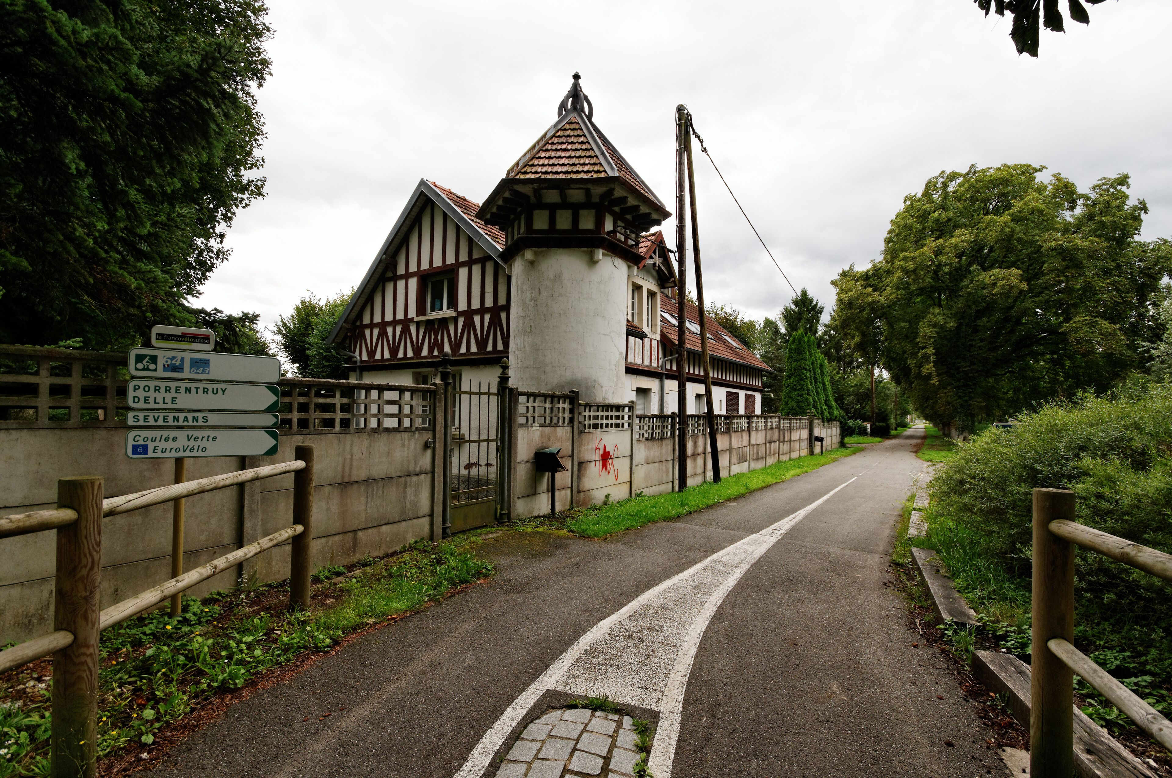 A traditional half-timbered building with a pointed roof, surrounded by greenery and a paved road.