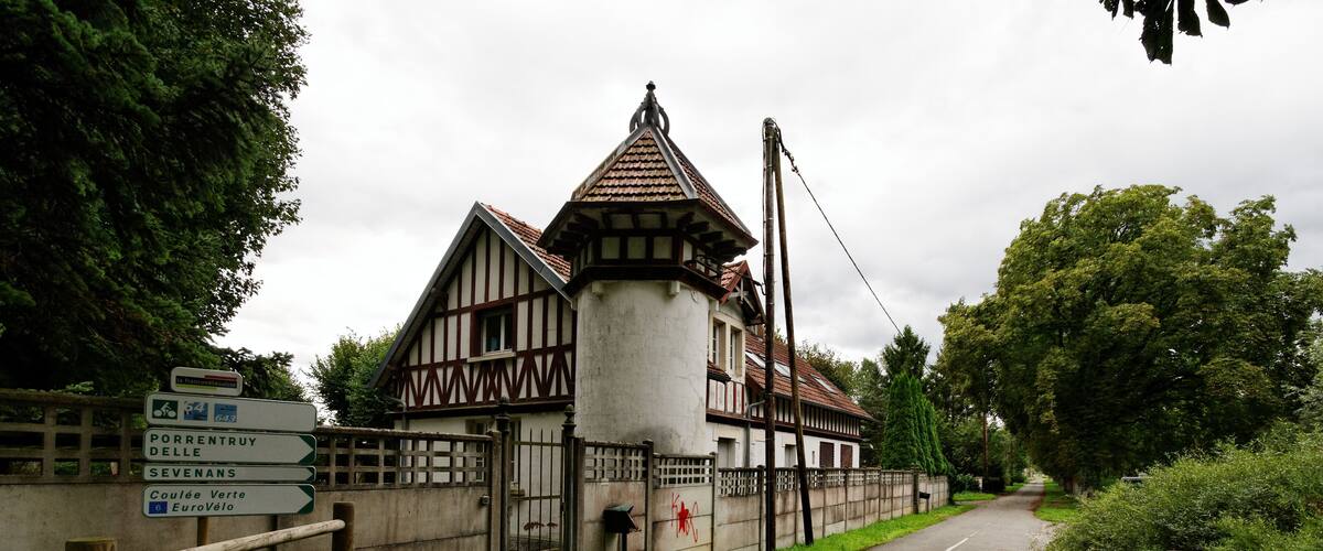 A traditional half-timbered building with a pointed roof, surrounded by greenery and a paved road.