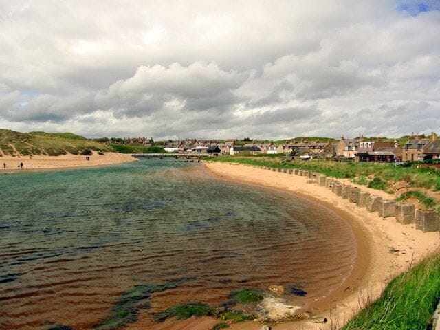 A coastal beach with clear water, sandy shore, and houses in the background.