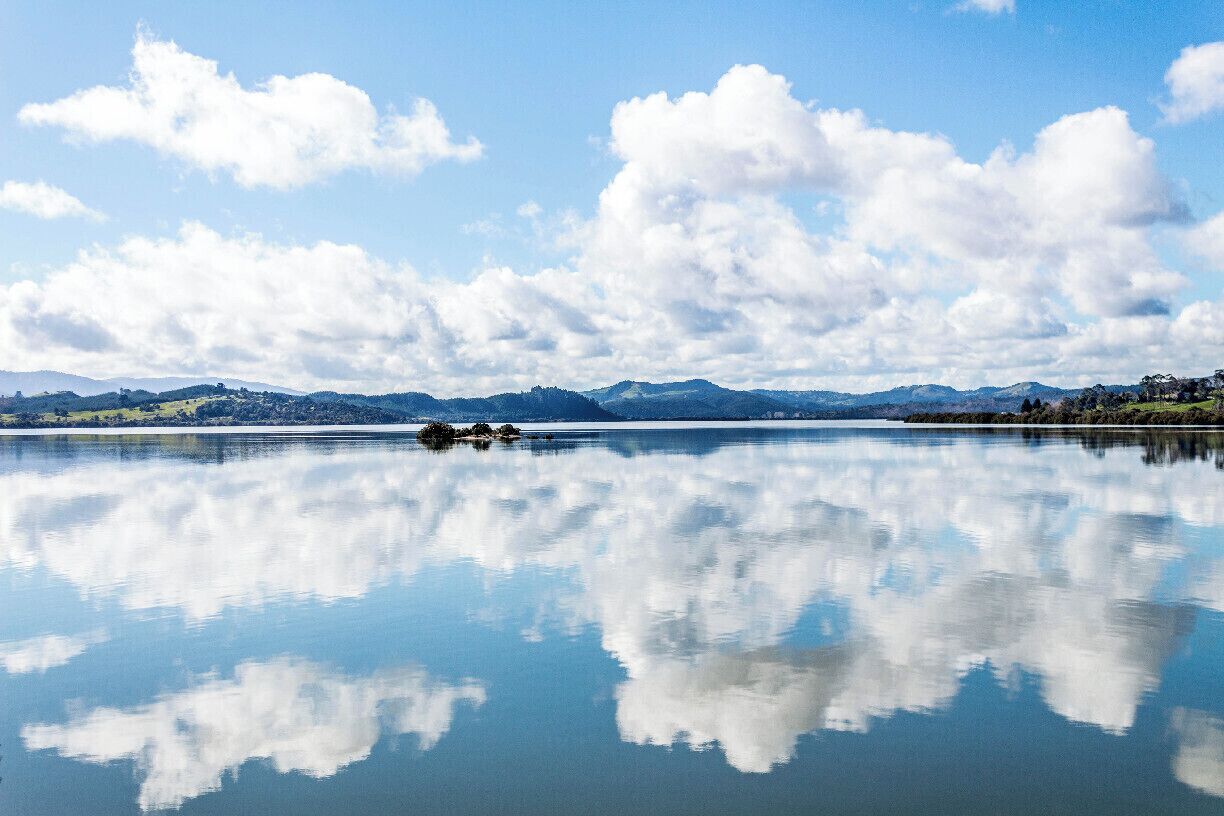 A calm lake with clear reflections of clouds, surrounded by distant mountains and a clear sky.