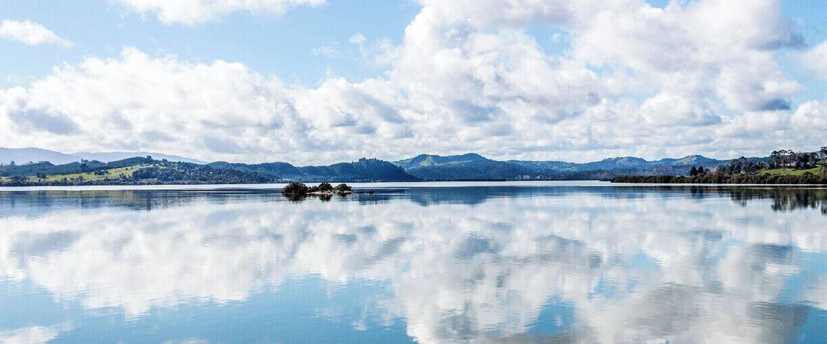 A calm lake with clear reflections of clouds, surrounded by distant mountains and a clear sky.
