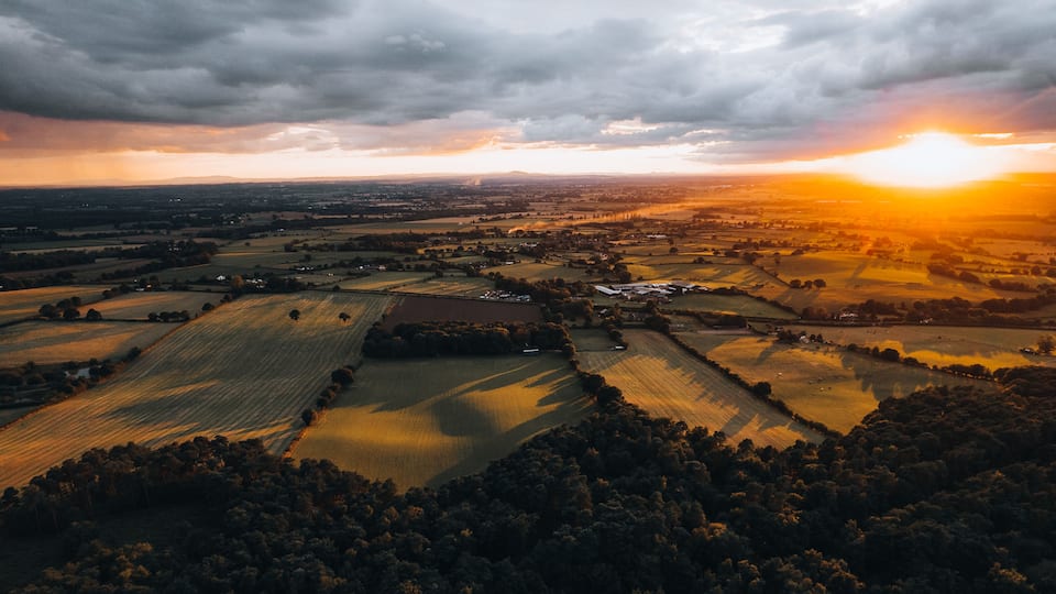 Een landelijk landschap met velden, bomen en een dorp bij zonsondergang.