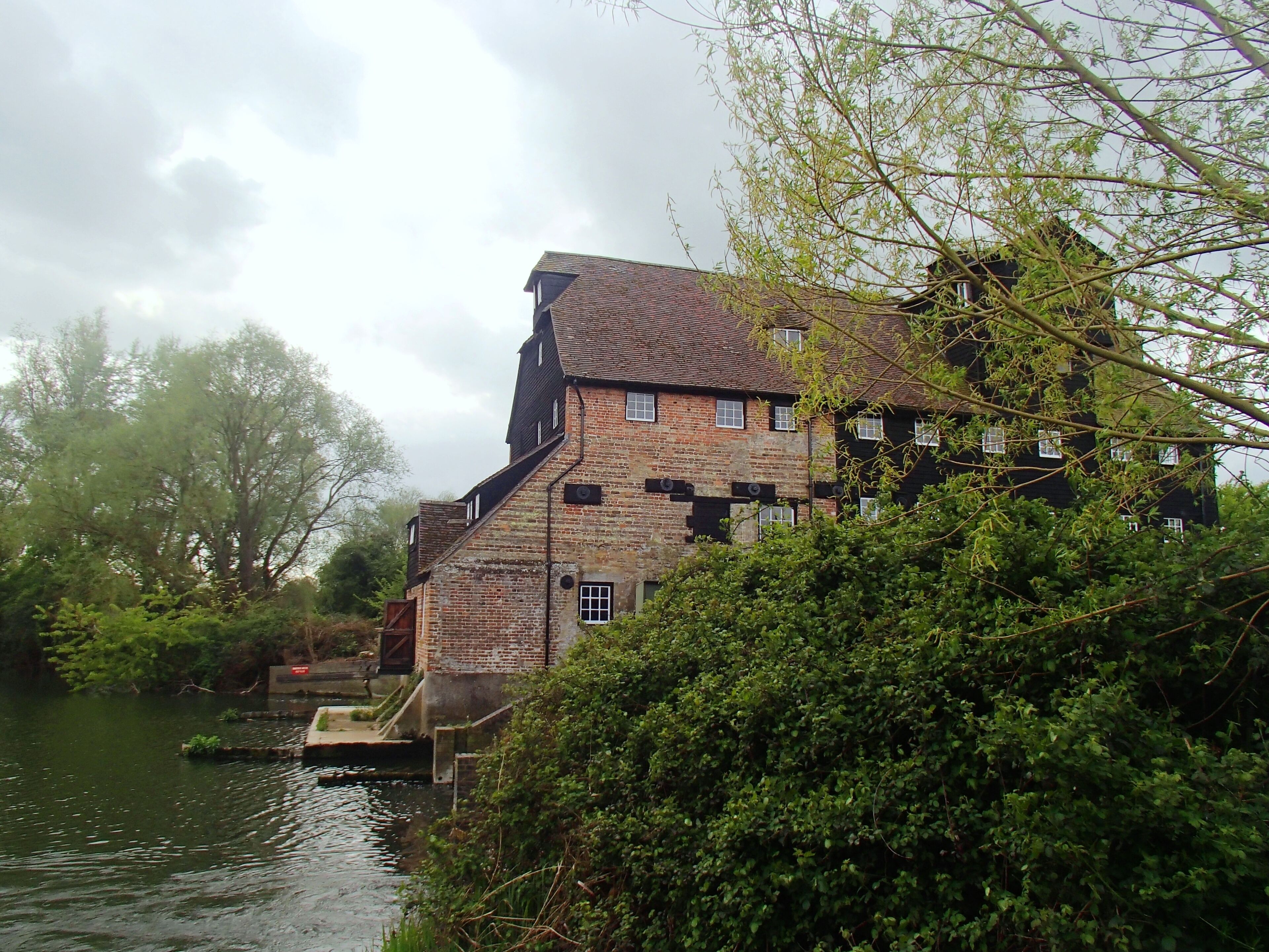 A historic brick building with a steep roof, situated by a river, surrounded by lush greenery.