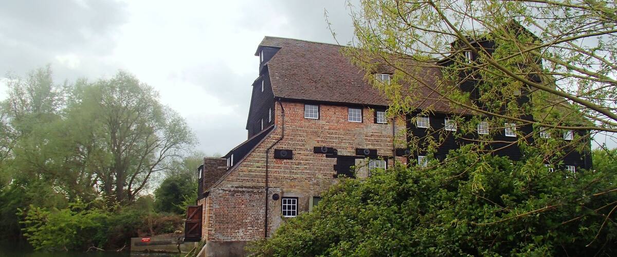 A historic brick building with a steep roof, situated by a river, surrounded by lush greenery.