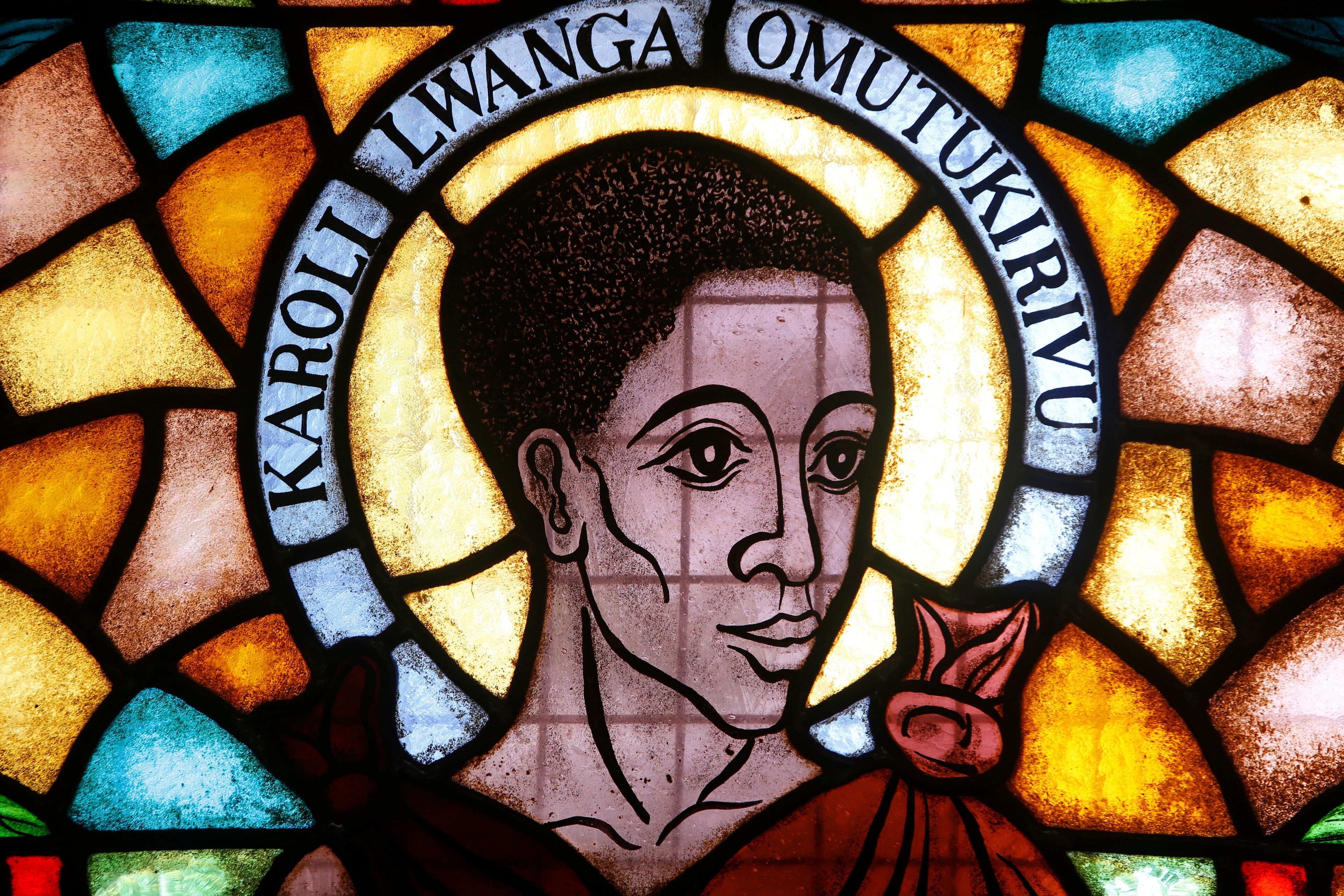 A stained glass window with a halo and the name "Karoli Lwanga Omukirivu".