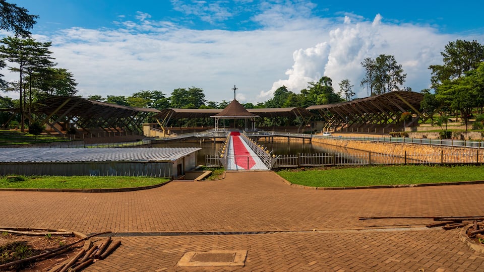 A covered walkway leading to a central structure with a cross on top, surrounded by greenery and a body of water.