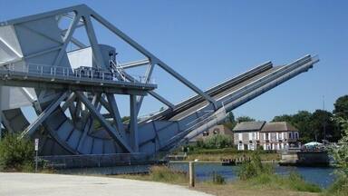 Une grande structure métallique formant un pont enjambant une rivière, avec une maison en arrière-plan.