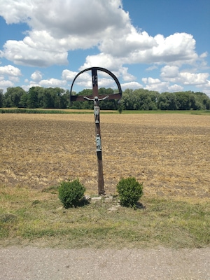 Una cruz de madera con inscripciones en un campo, con árboles y un cielo despejado al fondo.