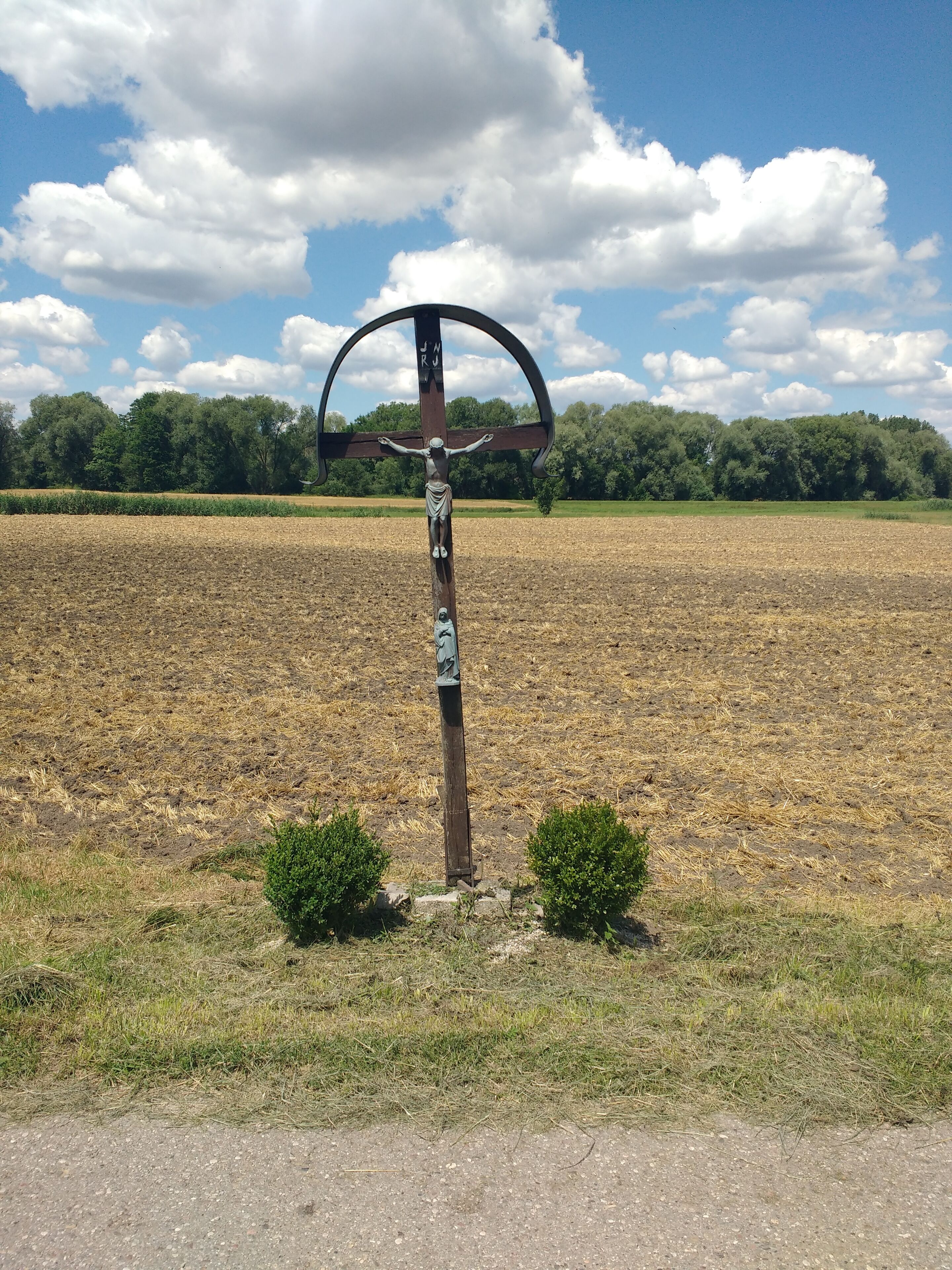 A wooden cross with inscriptions in a field with a backdrop of trees and a clear sky.