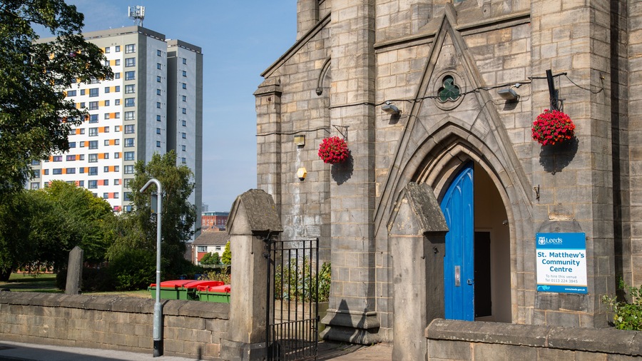 A historic stone building with a blue door, a sign for St. Matthew's Community Centre, and red flower baskets.