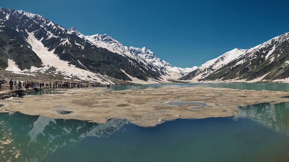 A lake surrounded by snow-capped mountains with a group of people on the left side.