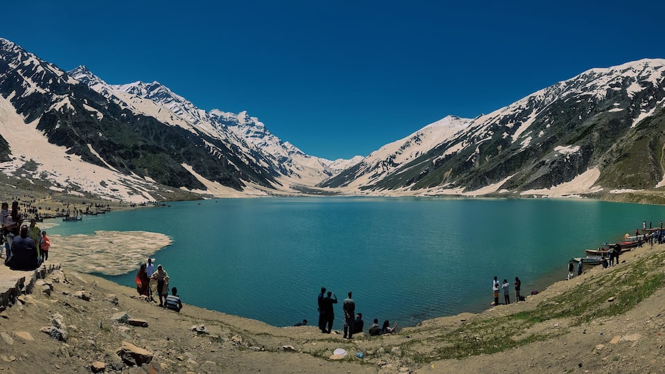 A lake surrounded by snow-capped mountains, with people walking and standing on the shore.
