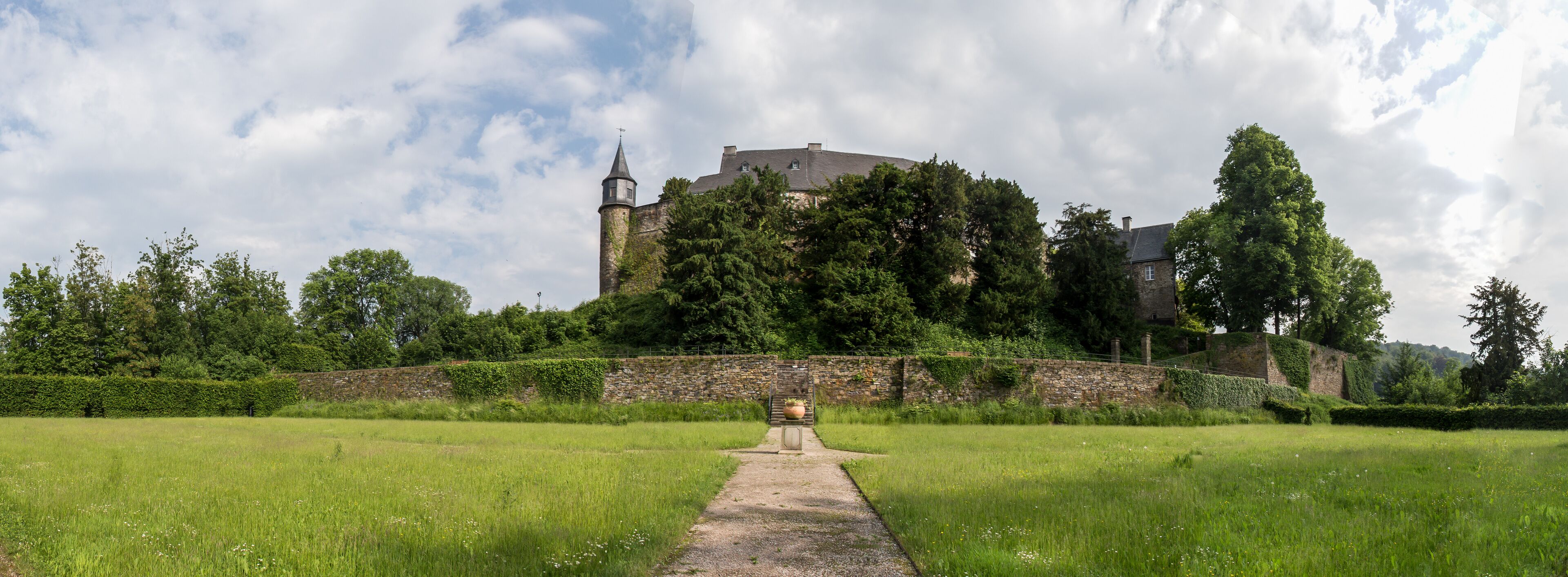 A castle on a hill with a pathway leading to it, surrounded by greenery and a stone wall.