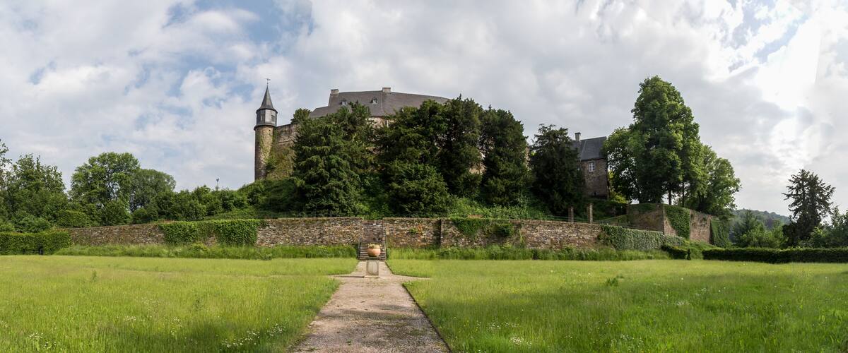 A castle on a hill with a pathway leading to it, surrounded by greenery and a stone wall.