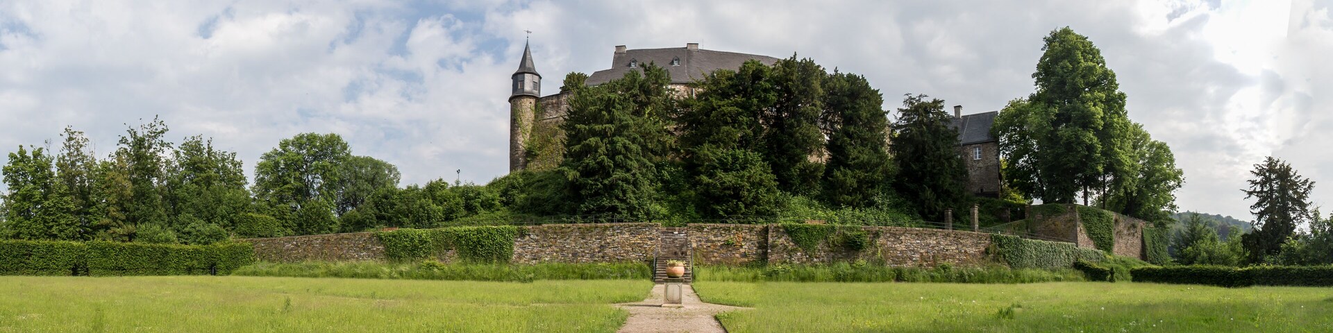 A castle on a hill with a pathway leading to it, surrounded by greenery and a stone wall.