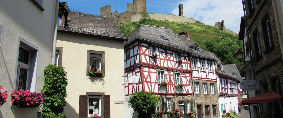 A cobblestone street lined with traditional half-timbered houses, a castle on a hill, and a sign for a local restaurant.