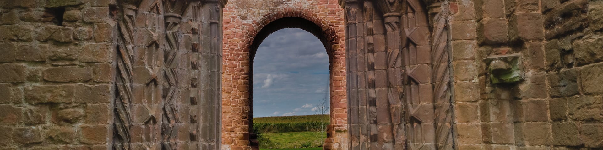 An arched stone doorway with intricate carvings, leading to a grassy field.