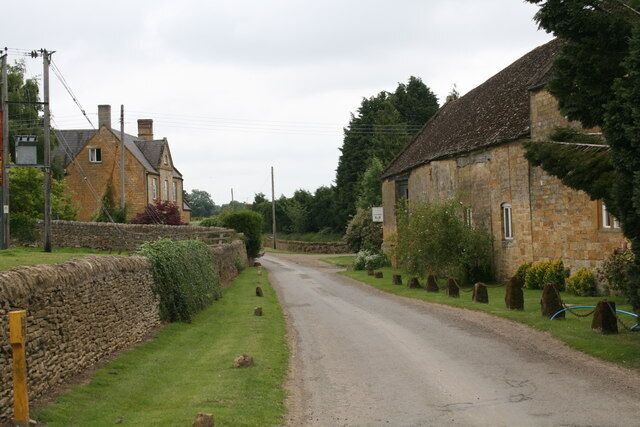 A rural road flanked by stone walls and houses, with a stone building on the right and a stone wall on the left.