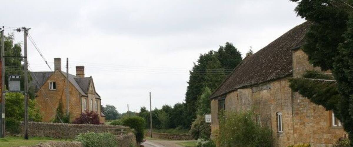 A rural road flanked by stone walls and houses, with a stone building on the right and a stone wall on the left.