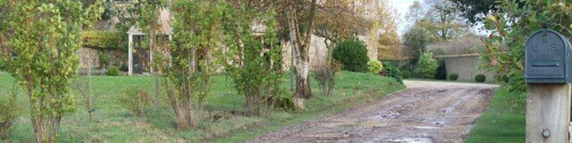 Uma estrada rural com uma caixa de correio, árvores e uma casa ao fundo.