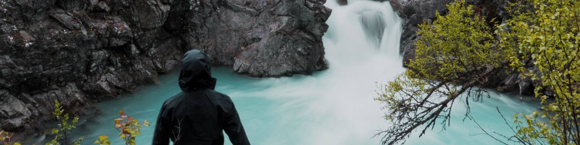 A person standing on a rocky outcrop overlooking a turquoise waterfall in a forested valley with mountains in the background.